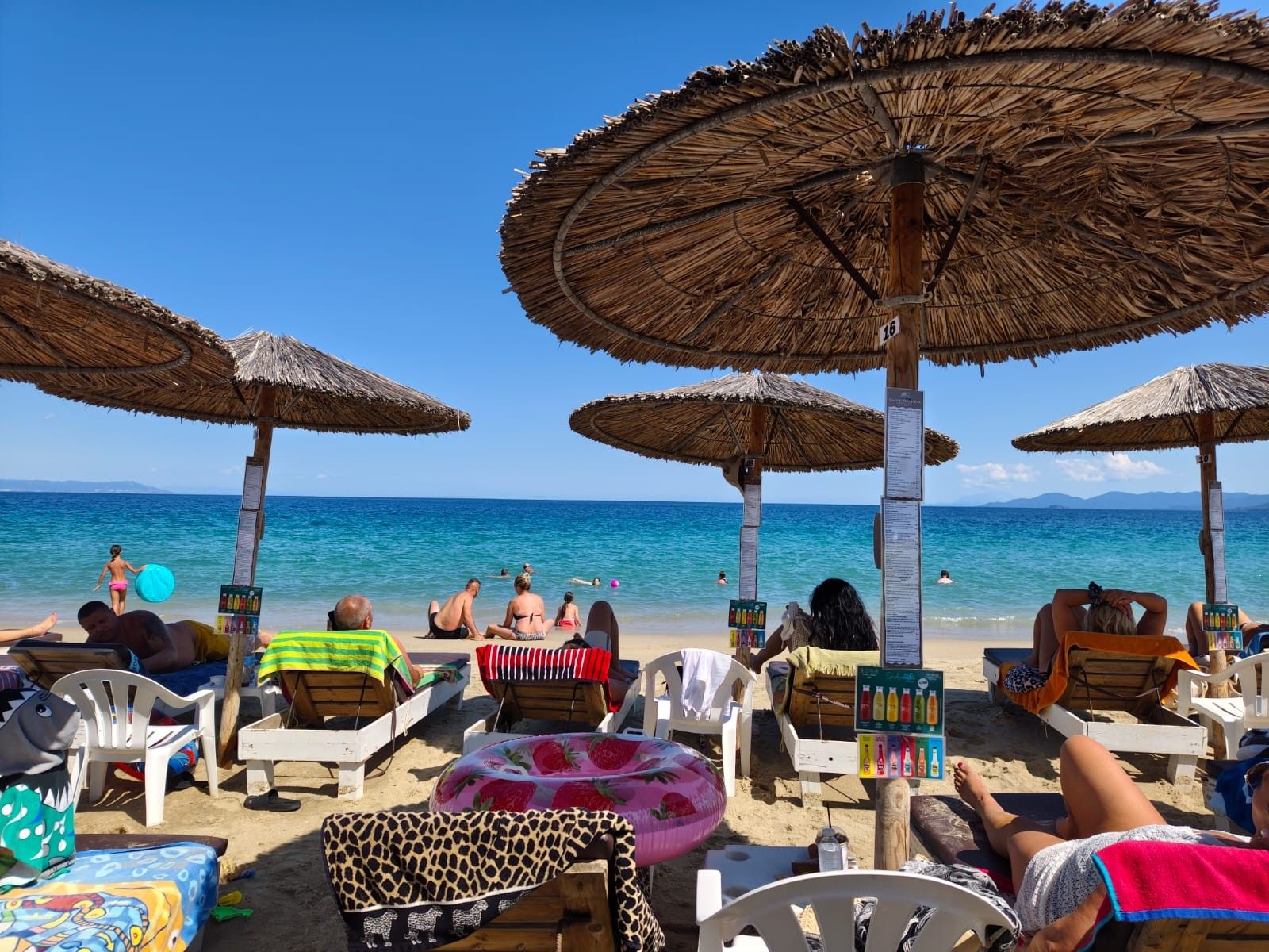 A beach with sun loungers and thatched umbrellas, a clear blue sea, and a distant mountain range.