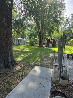 A fenced outdoor area with a concrete path, a large tree, and two small buildings. - Dancin' Dogs, The Dog House, is a one bedroom bungalow in Newberry FL (Newberry)