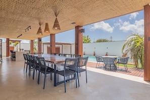 Un área de comedor al aire libre con vista a la piscina, que incluye una mesa grande y sillas, además de un espacio de estar cubierto con luces colgantes.
