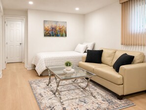 Een moderne woonkamer met een glazen salontafel, een beige bank, een bed met een geruit dekbed en een schilderij aan de muur.