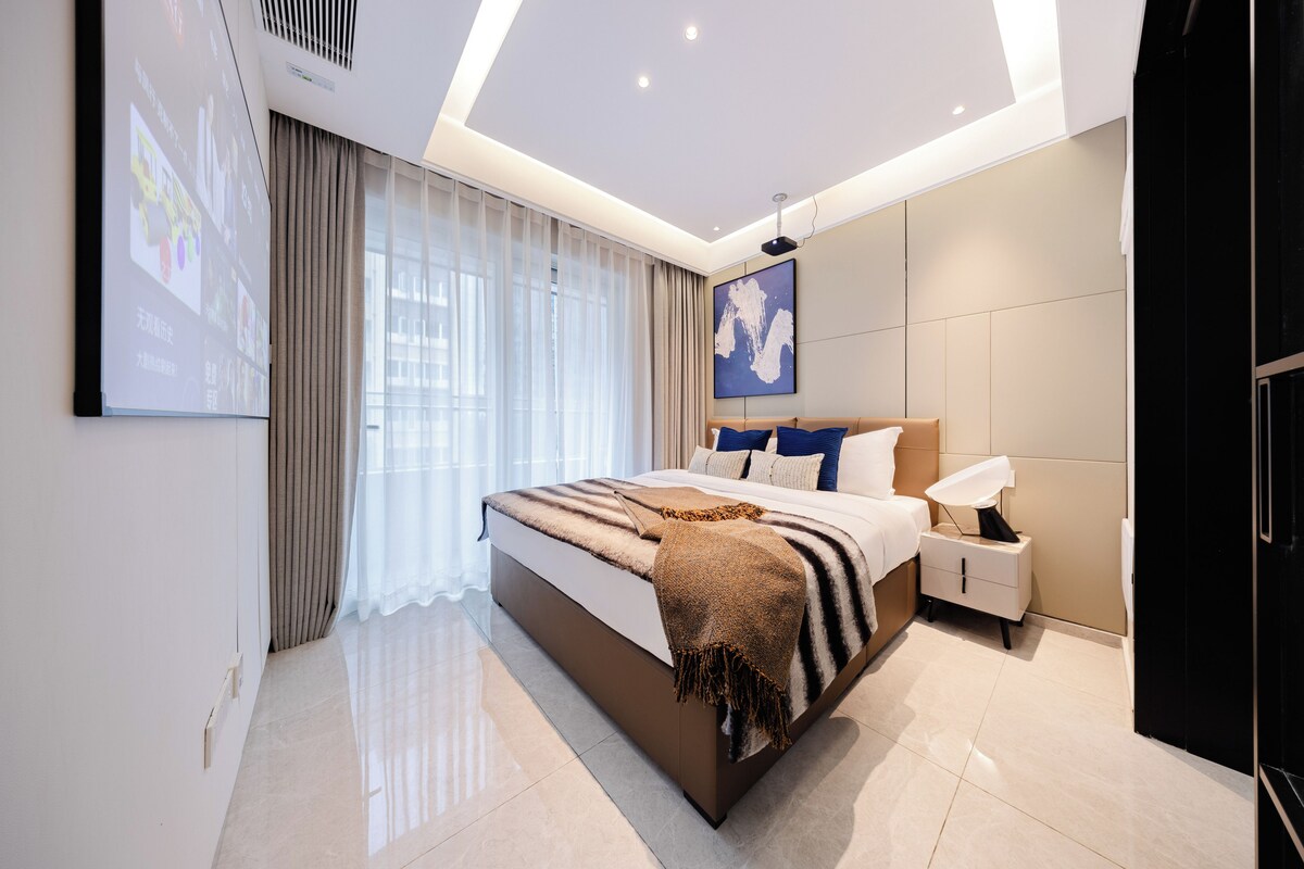 A modern bedroom with a large bed, bedside tables, a projector screen, and a ceiling light.