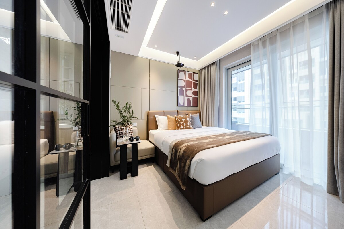 A modern hotel room with a large bed, a bedside table, a small table with a plant, and a large window with curtains.