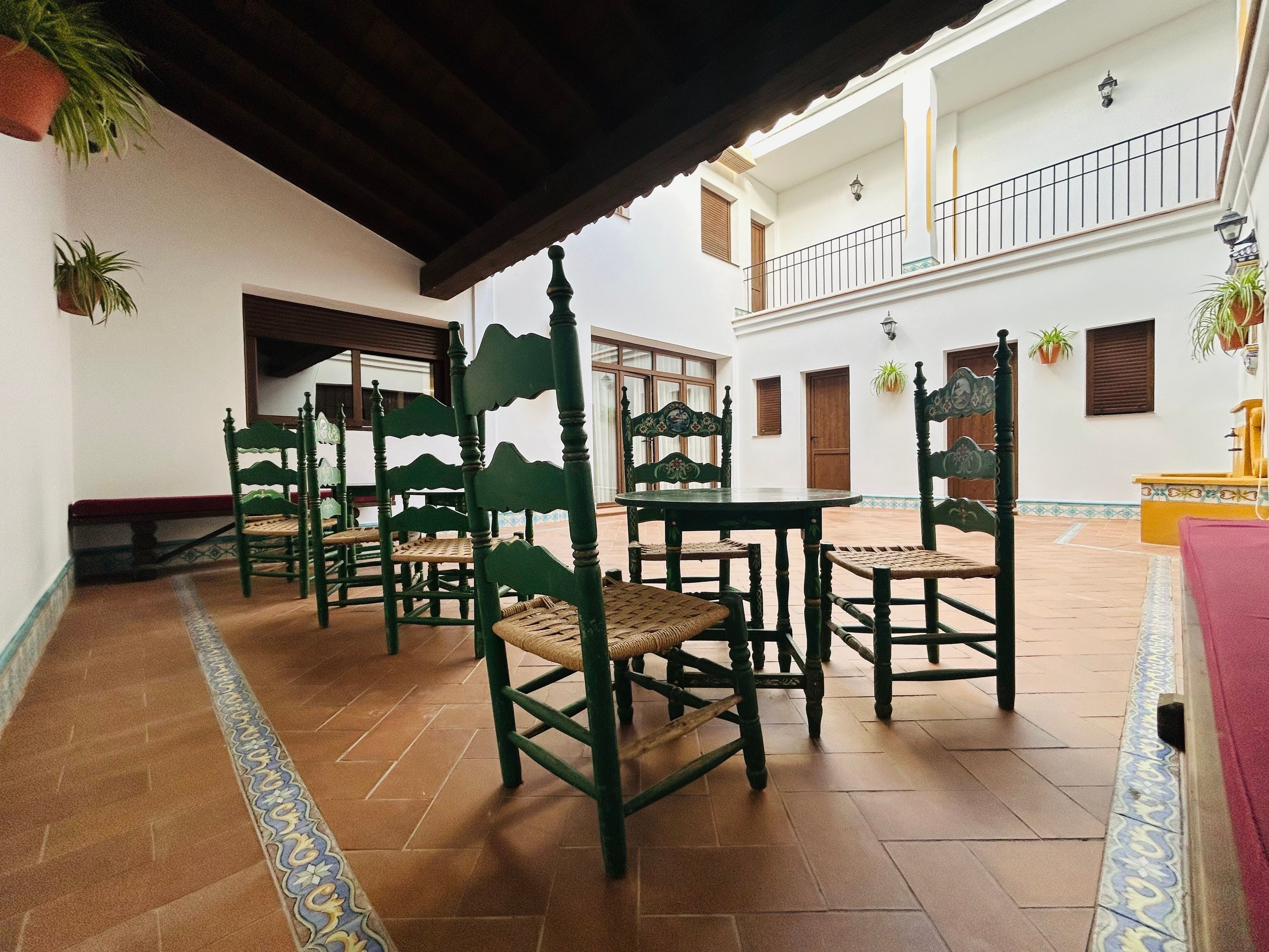 A courtyard with tiled flooring, green wooden chairs, a bench, and a yellow table.