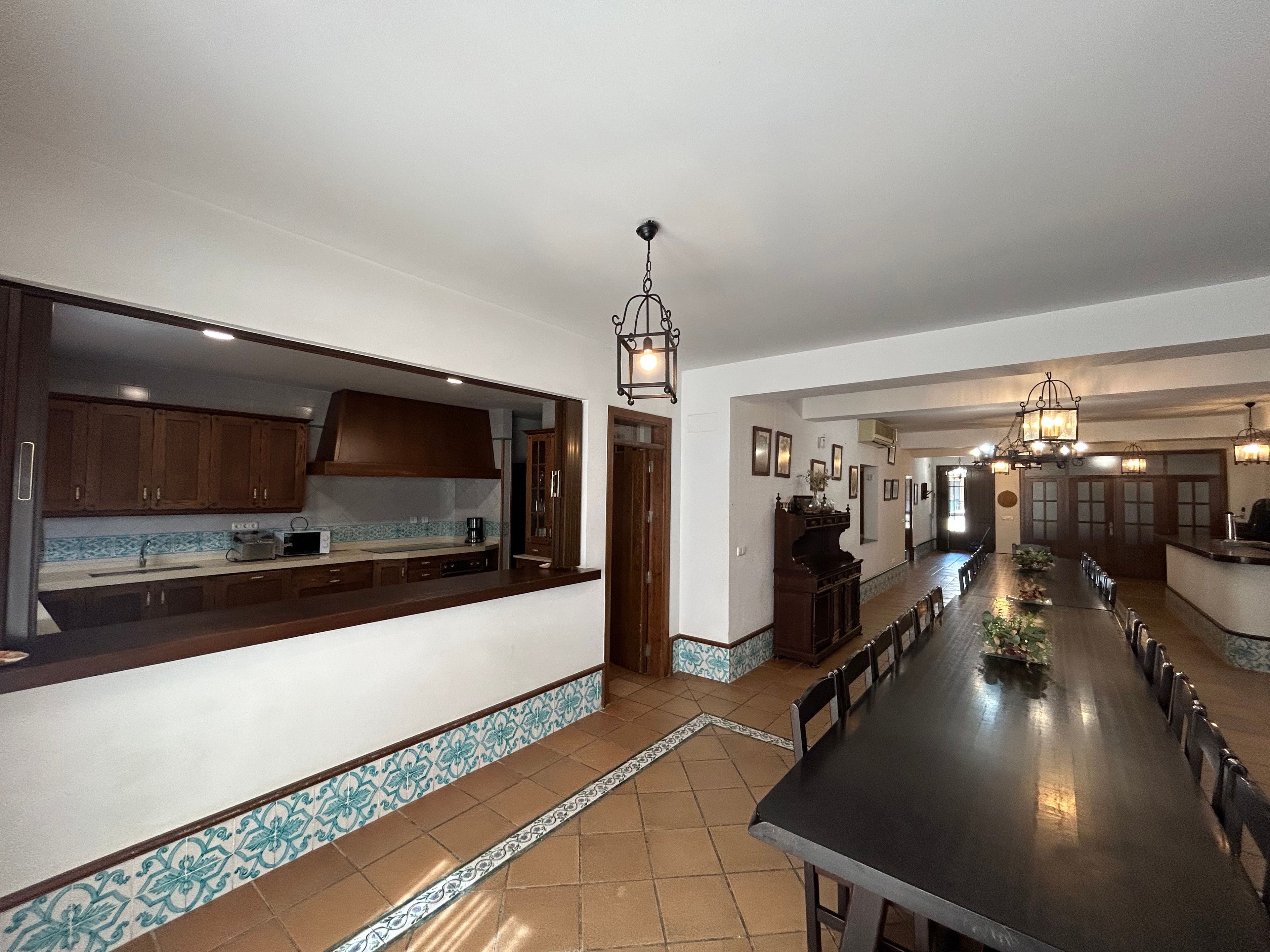 A long dining table with chairs, a kitchen area with wooden cabinets, and a hallway with a chandelier.