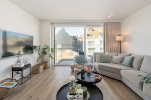 A modern living room with a sofa, a coffee table, and a television. There is a balcony with a view of buildings. - 4-Beaufort - everything within walking distance (Nieuwpoort)