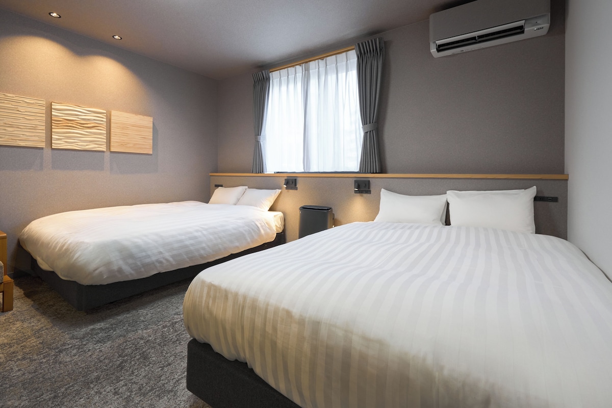 A hotel room with two beds, a window with curtains, an air conditioning unit, and a wall-mounted light fixture.