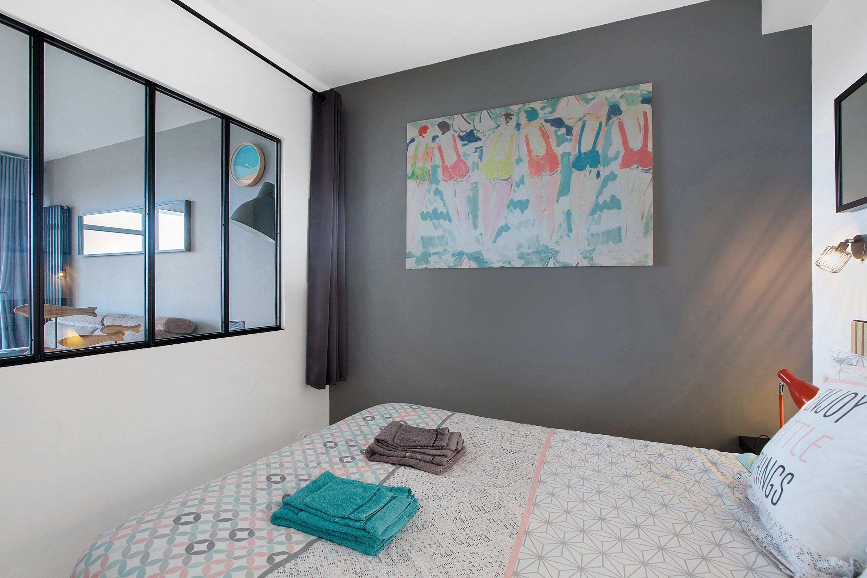 Une chambre à coucher moderne avec un grand lit, une fenêtre avec des stores, une horloge murale et un tableau abstrait aux couleurs vives.