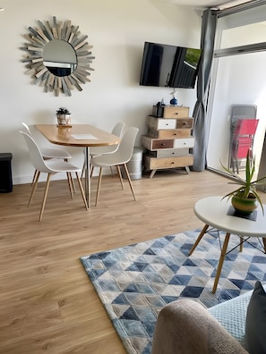 Una sala de estar moderna con comedor, televisor montado en la pared y una planta en una mesita.