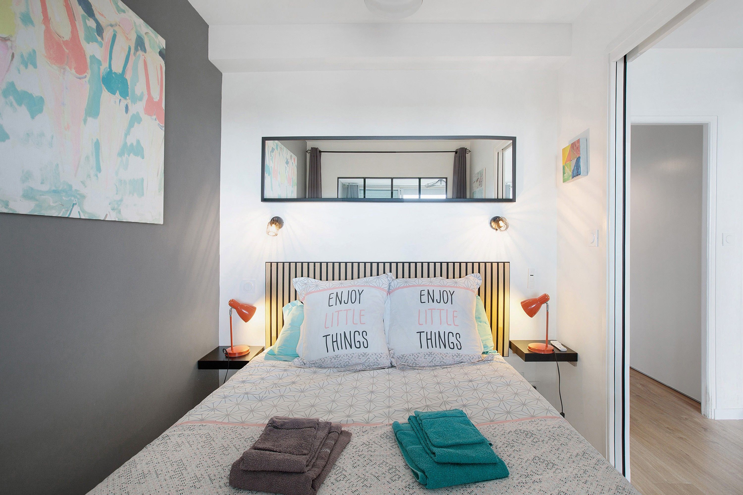 A bedroom with a bed, two bedside tables, a nightstand, a mirror, and a wall with abstract art.