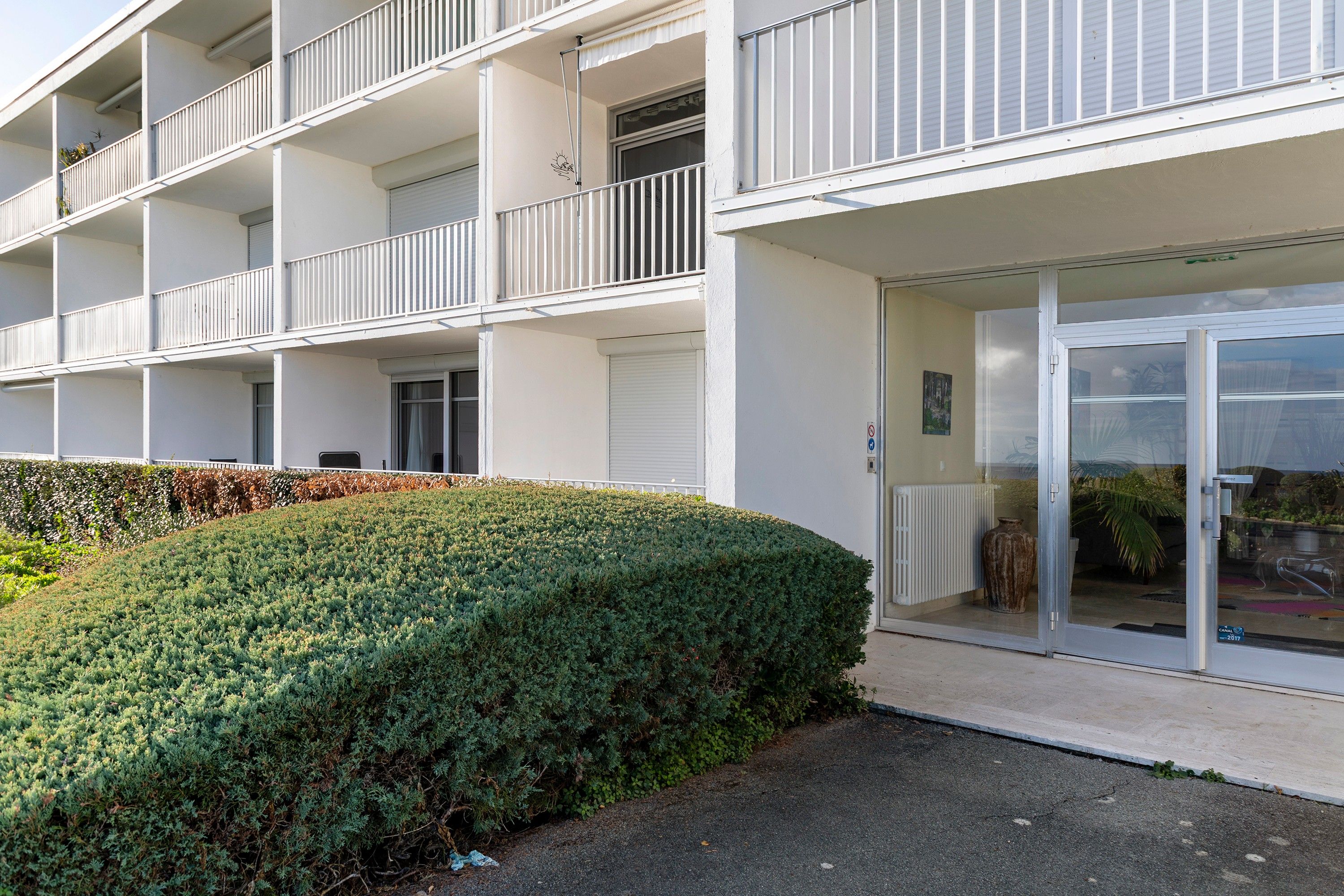 Un immeuble à plusieurs étages, doté de balcons et d’une haie bien entretenue.