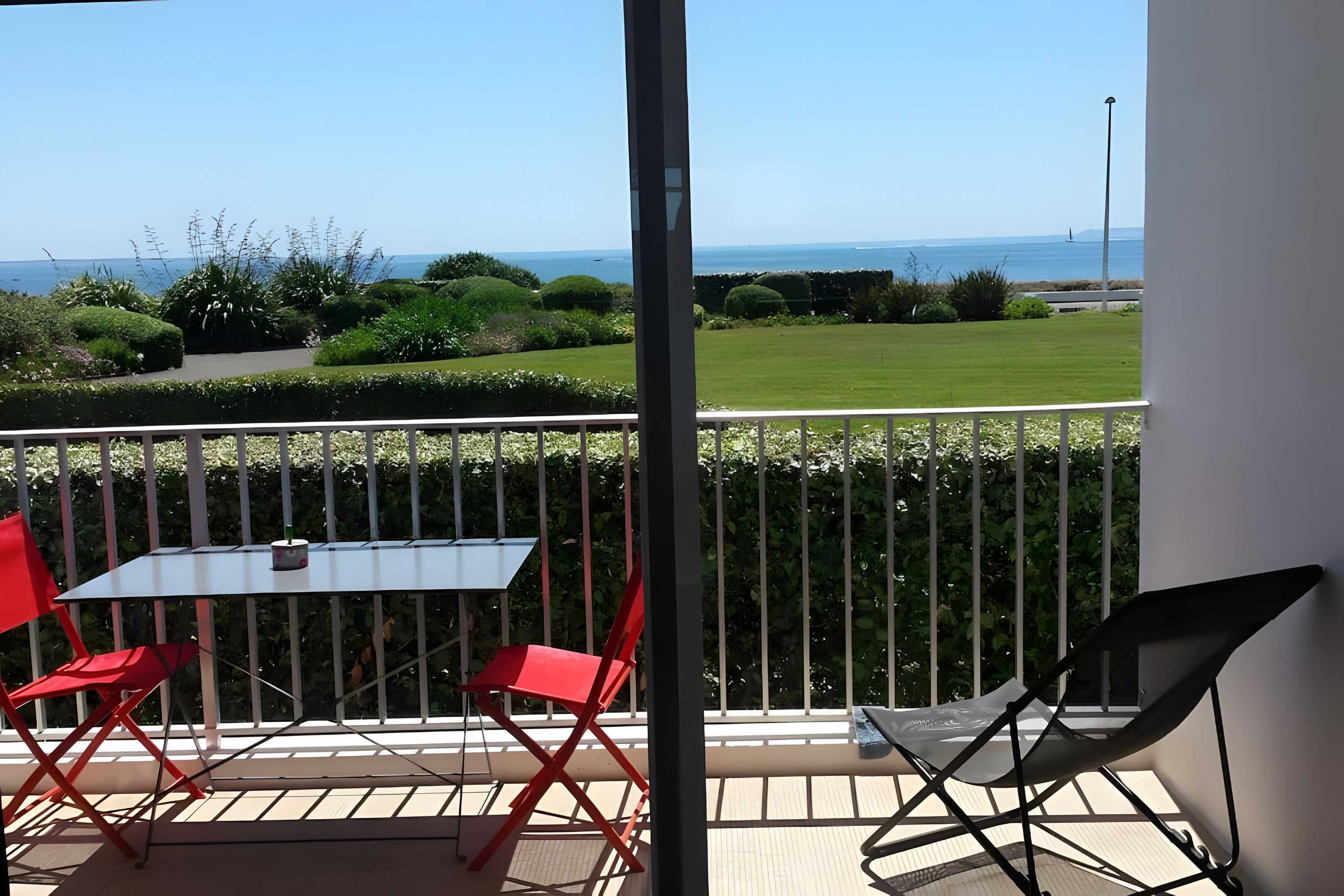 A balcony with a table and chairs, a view of the sea, and a well-maintained garden.