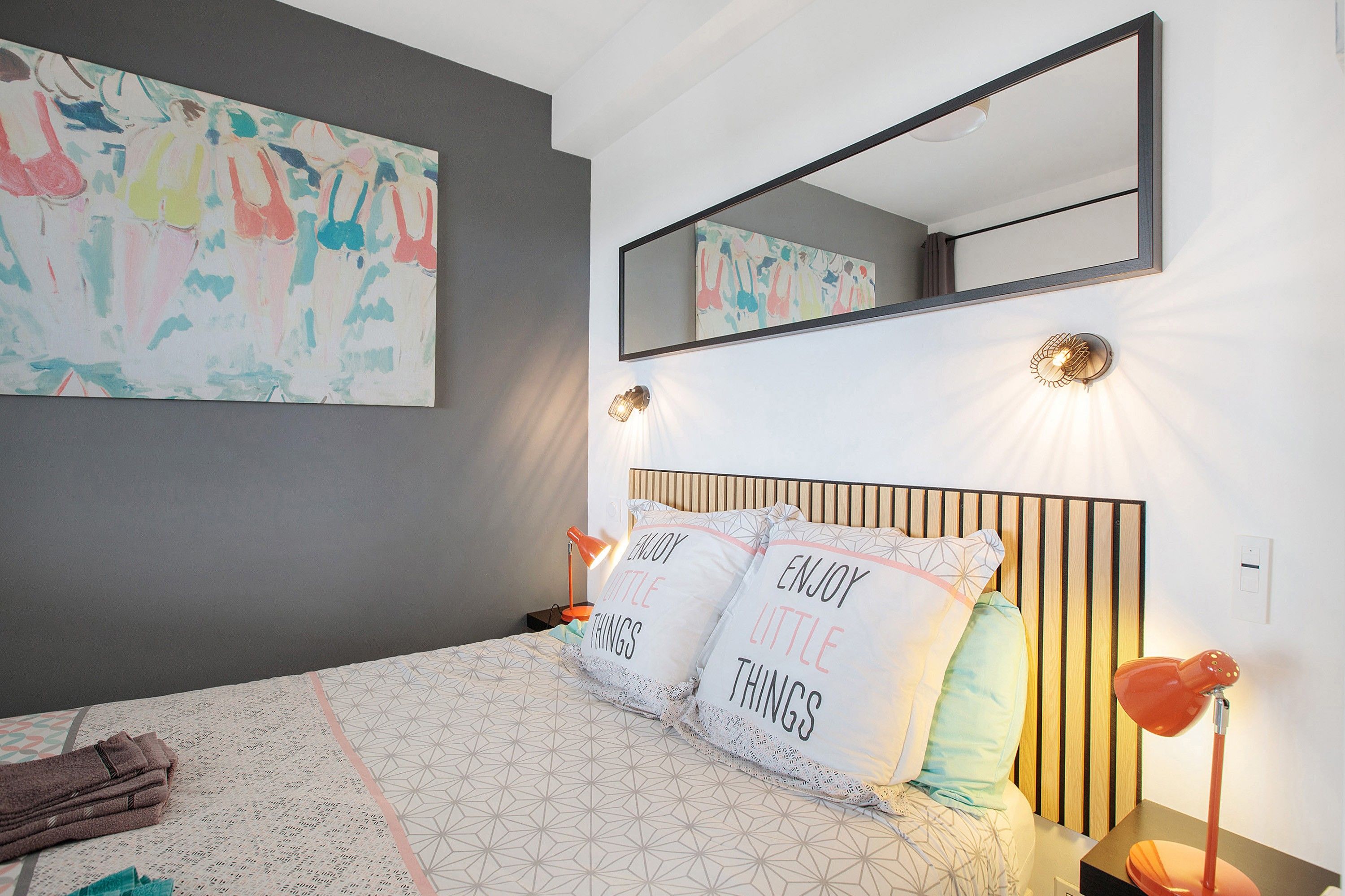Une chambre à coucher avec un lit, des oreillers, une table de chevet, une lampe, un miroir fixé au mur et des œuvres d’art abstraites.