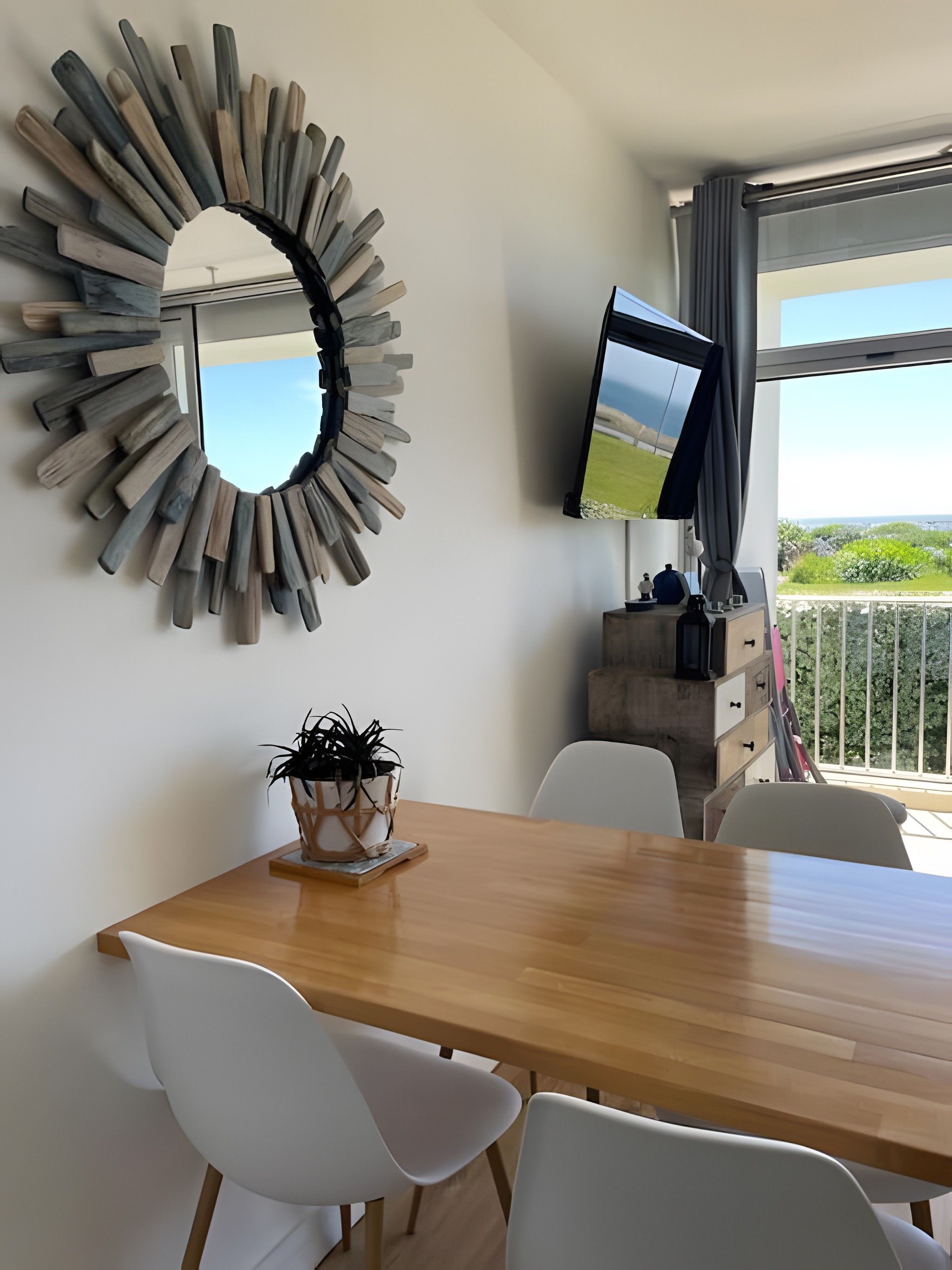 Een eethoek met een houten tafel, witte stoelen, een plant in pot, een televisie op een standaard en uitzicht op groen door een raam.