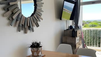 Een eethoek met een houten tafel, witte stoelen, een plant in pot, een televisie op een standaard en uitzicht op groen door een raam.