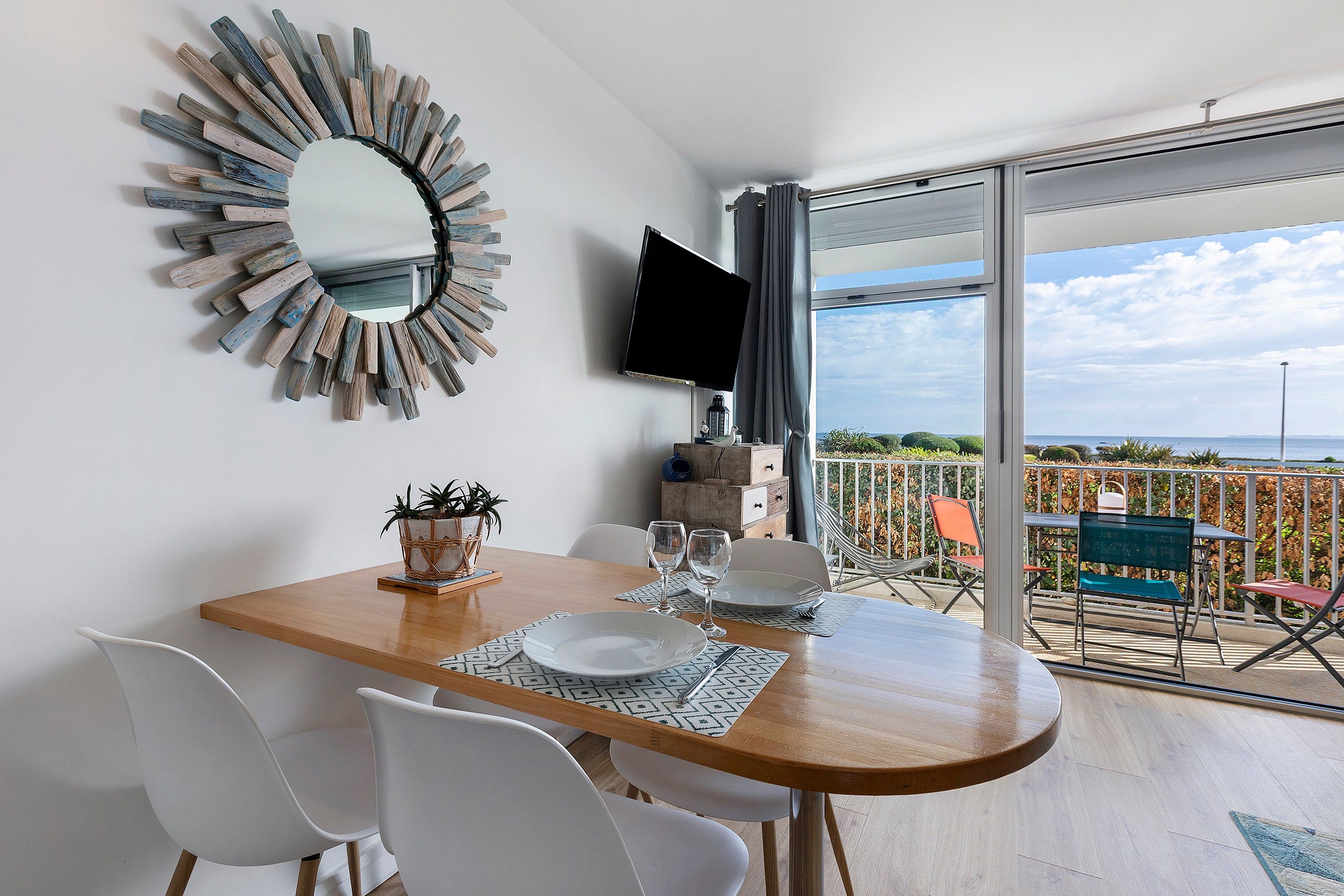 Un coin repas avec une table en bois, des chaises blanches, un miroir décoratif et un téléviseur à écran plat.