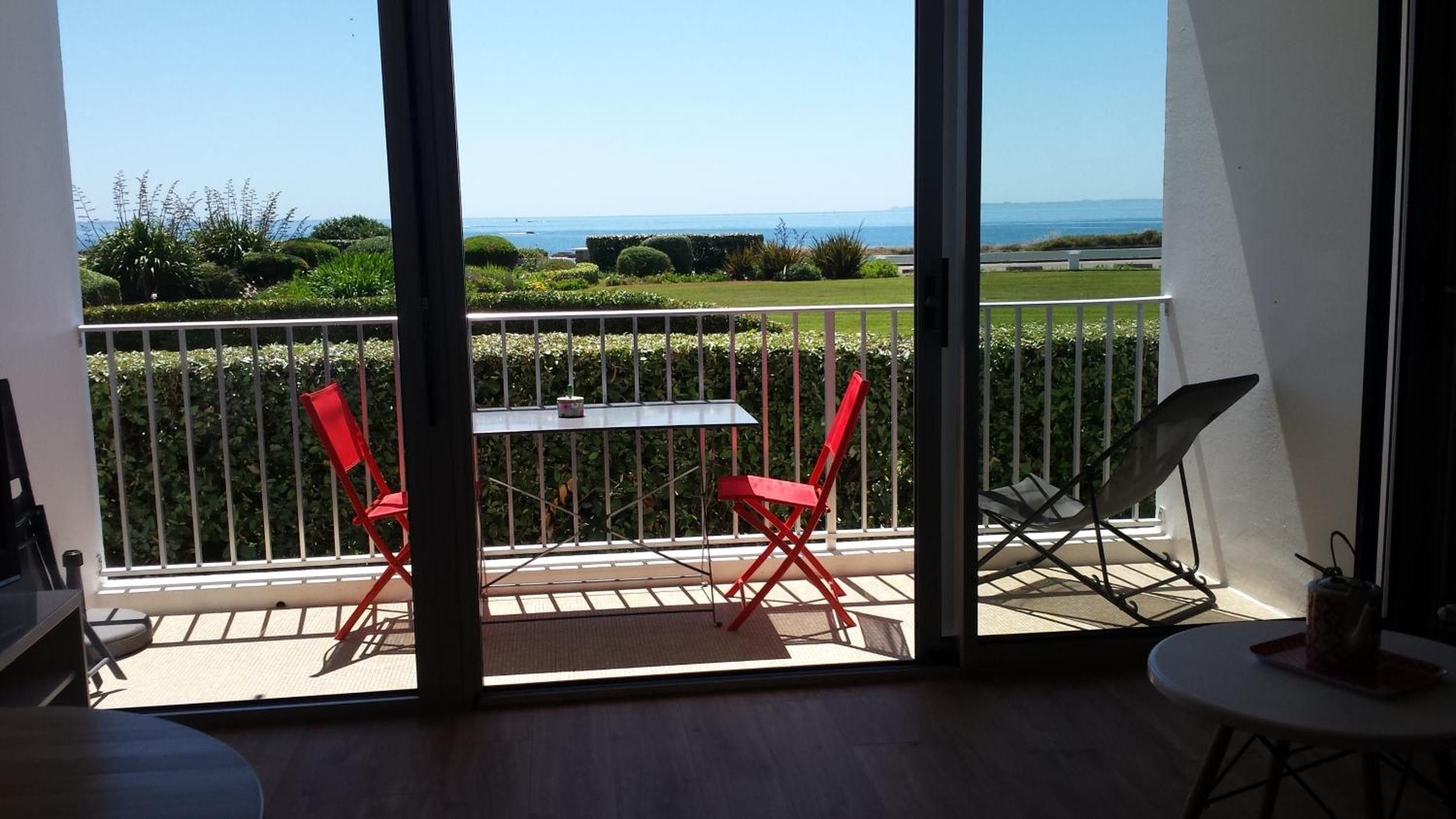 A balcony with a table and two red chairs, overlooking a well-maintained garden and the sea.