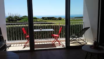 A balcony with a table and two red chairs, overlooking a well-maintained garden and the sea.