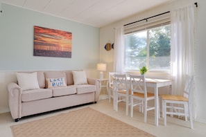 A living room with a beige sofa, a dining table with chairs, a window with curtains, and a painting on the wall. - Beach Access! Sunrise Side Resort Retreat (Greenbush)