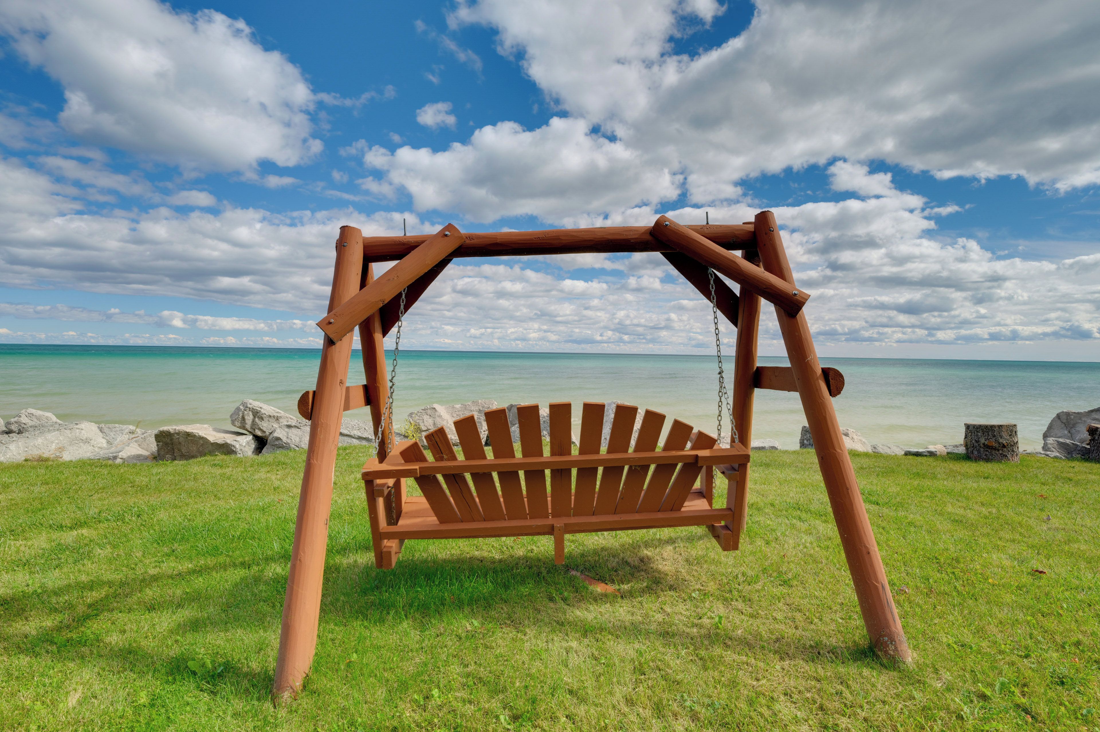 A wooden swing set with a bench seat, located on a grassy area with a view of a calm sea and a partly cloudy sky.