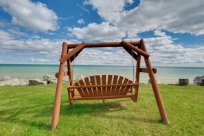 A wooden swing set with a bench seat, located on a grassy area with a view of a calm sea and a partly cloudy sky. - Waterfront Lake Huron Condo at Sunrise Side Resort (Greenbush)