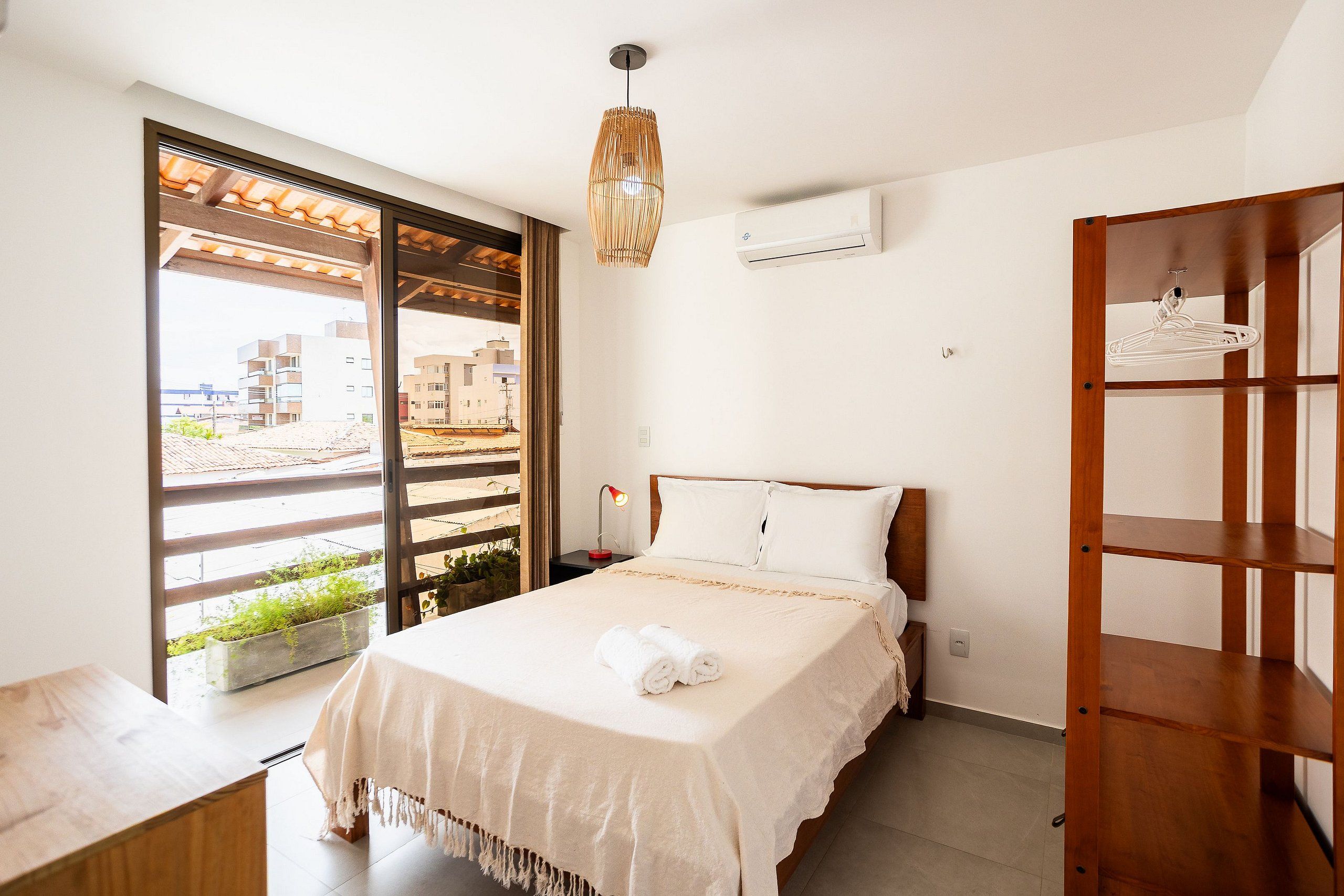 A bedroom with a bed, a bedside lamp, a hanging light fixture, a window with a view, and an air conditioning unit.