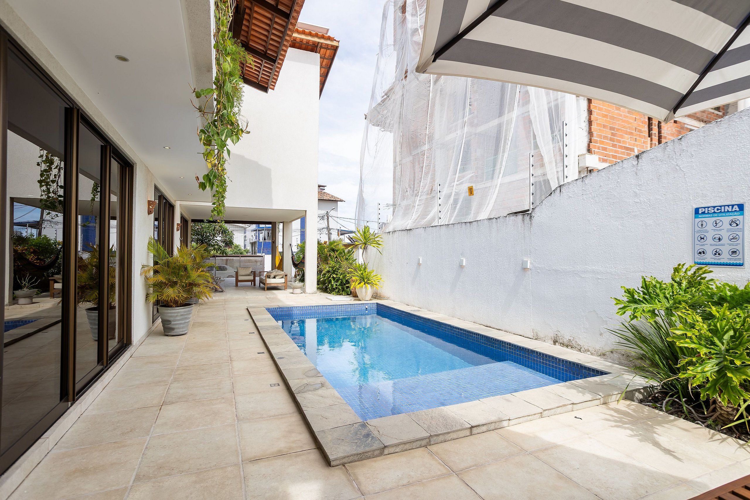 A swimming pool with a tiled edge, surrounded by a patio area with potted plants and a covered seating area.