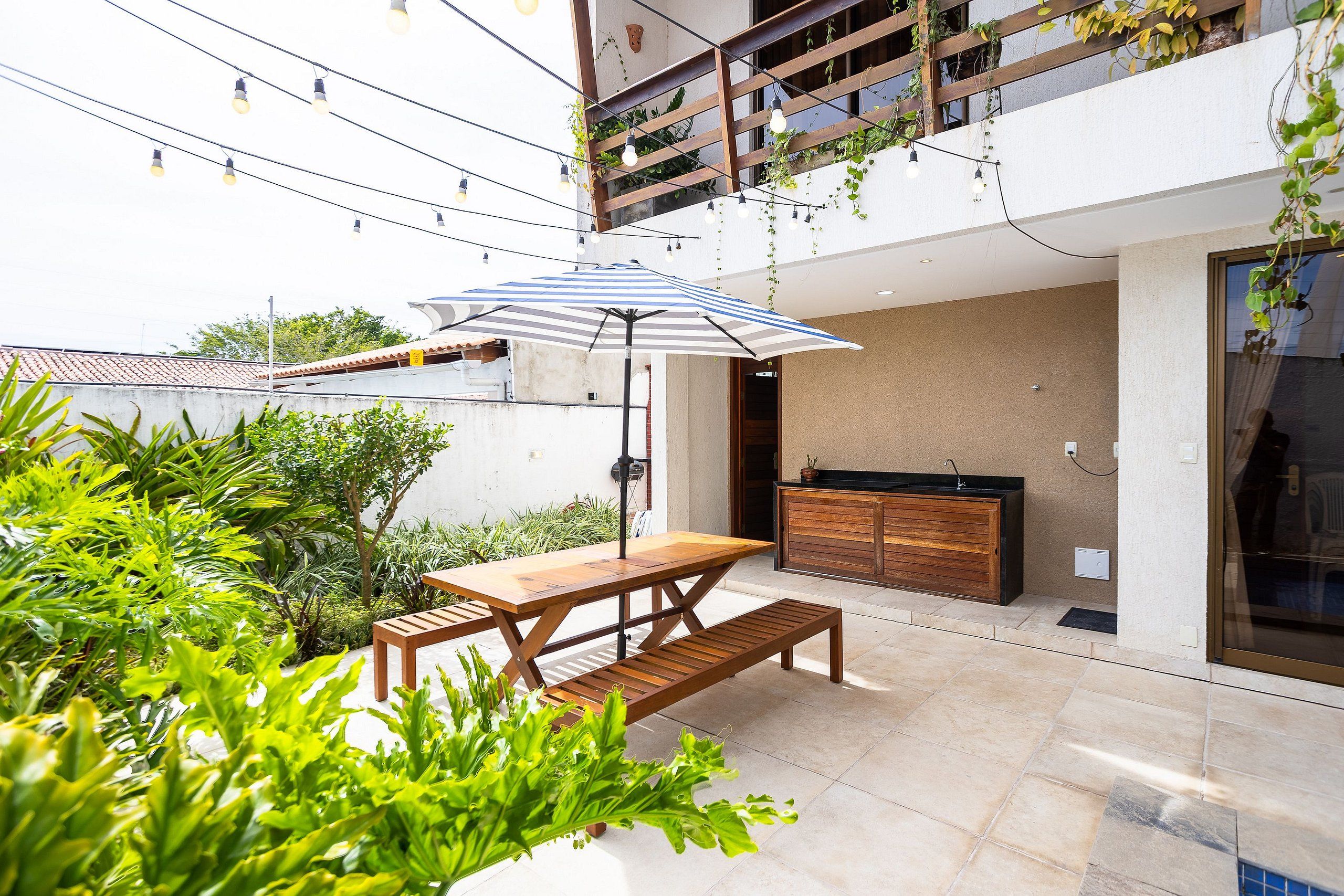 An outdoor patio with a wooden table and bench, surrounded by lush greenery.