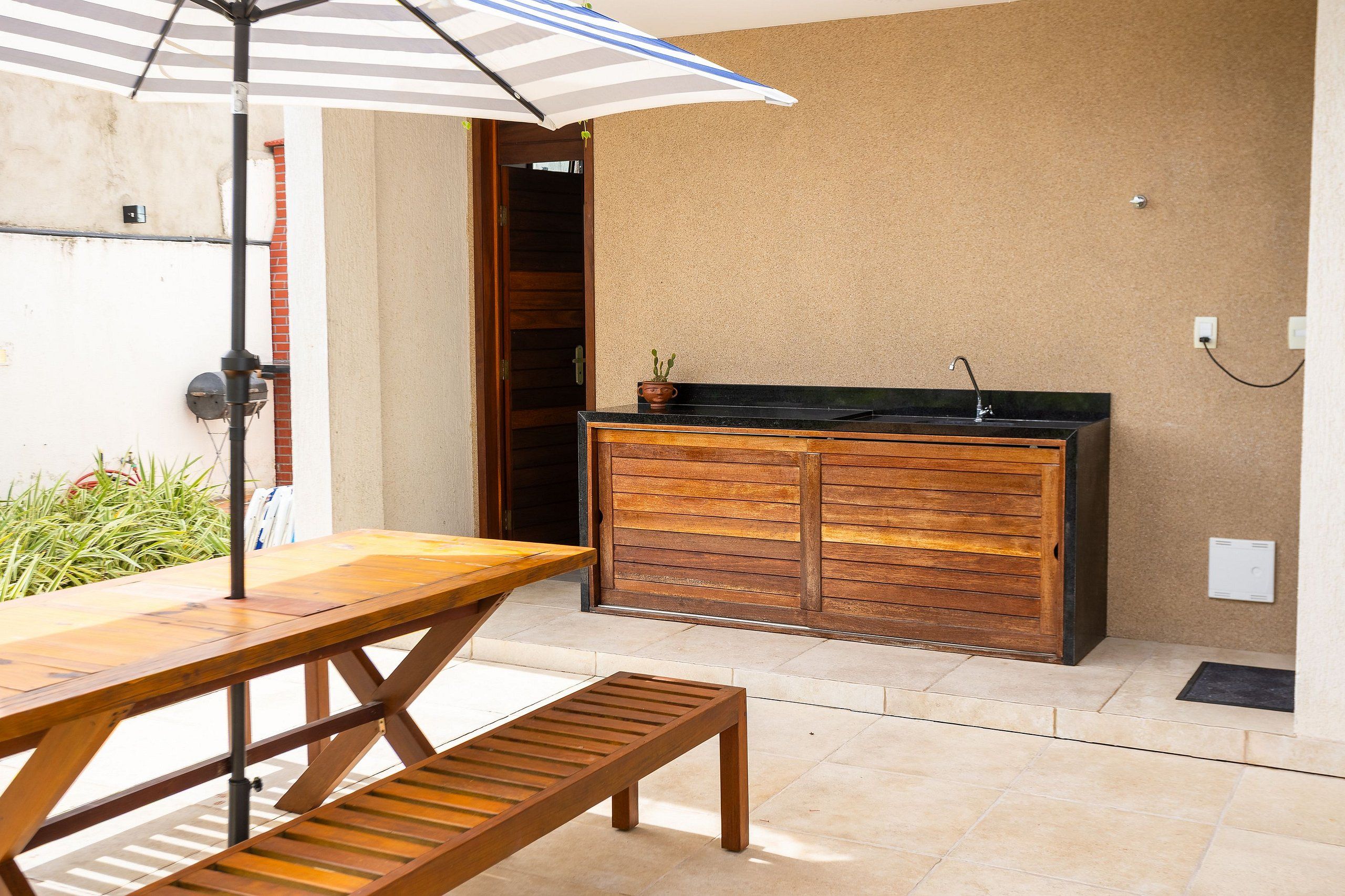 Eine Terrasse im Freien mit einem Holztisch und einer Bank, einem gestreiften Sonnenschirm und einer integrierten Küchenzeile mit Spüle und Arbeitsplatte.