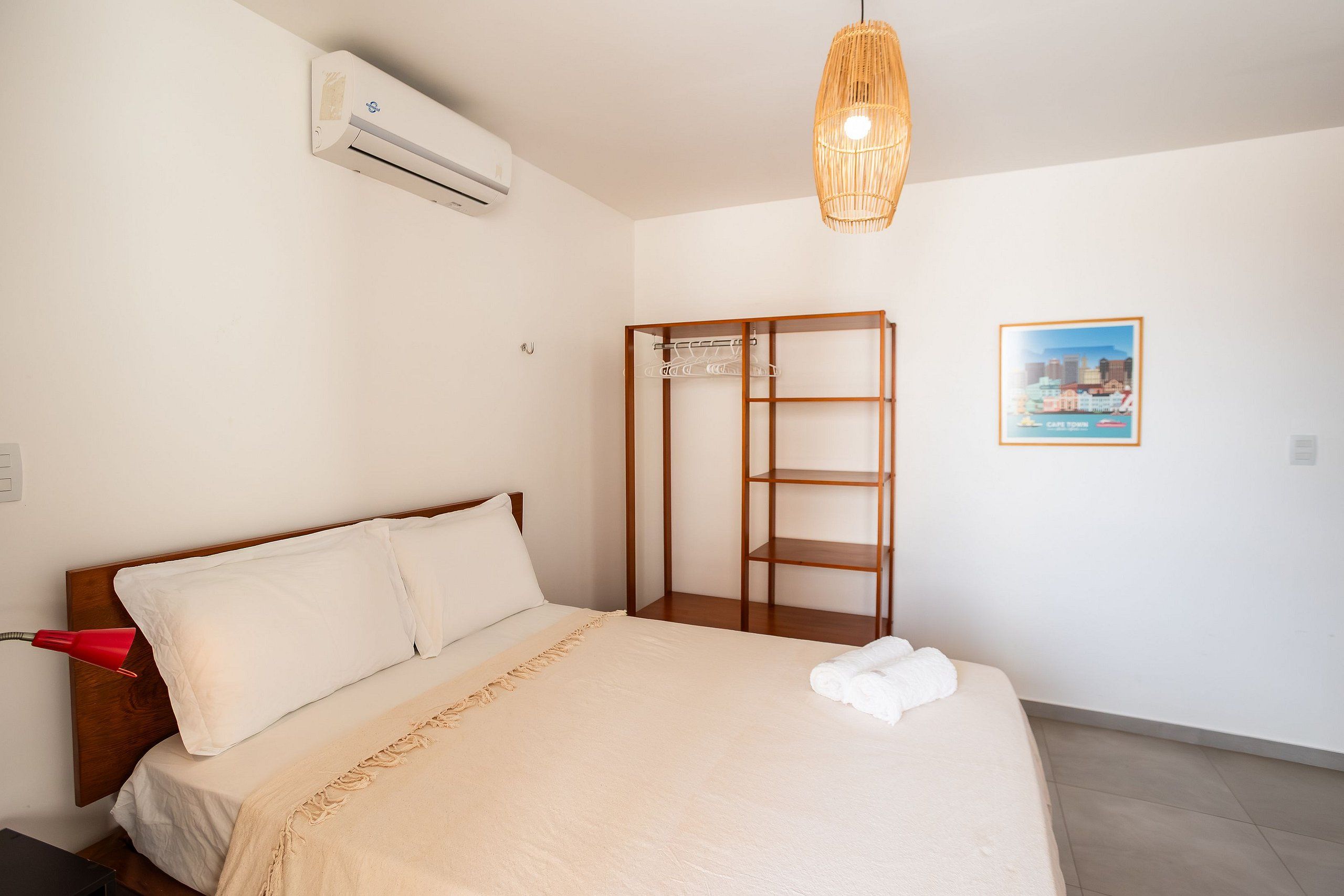 Une chambre d’hôtel avec un lit, une armoire, un appareil de climatisation, un luminaire suspendu et une affiche représentant un paysage urbain accrochée au mur.