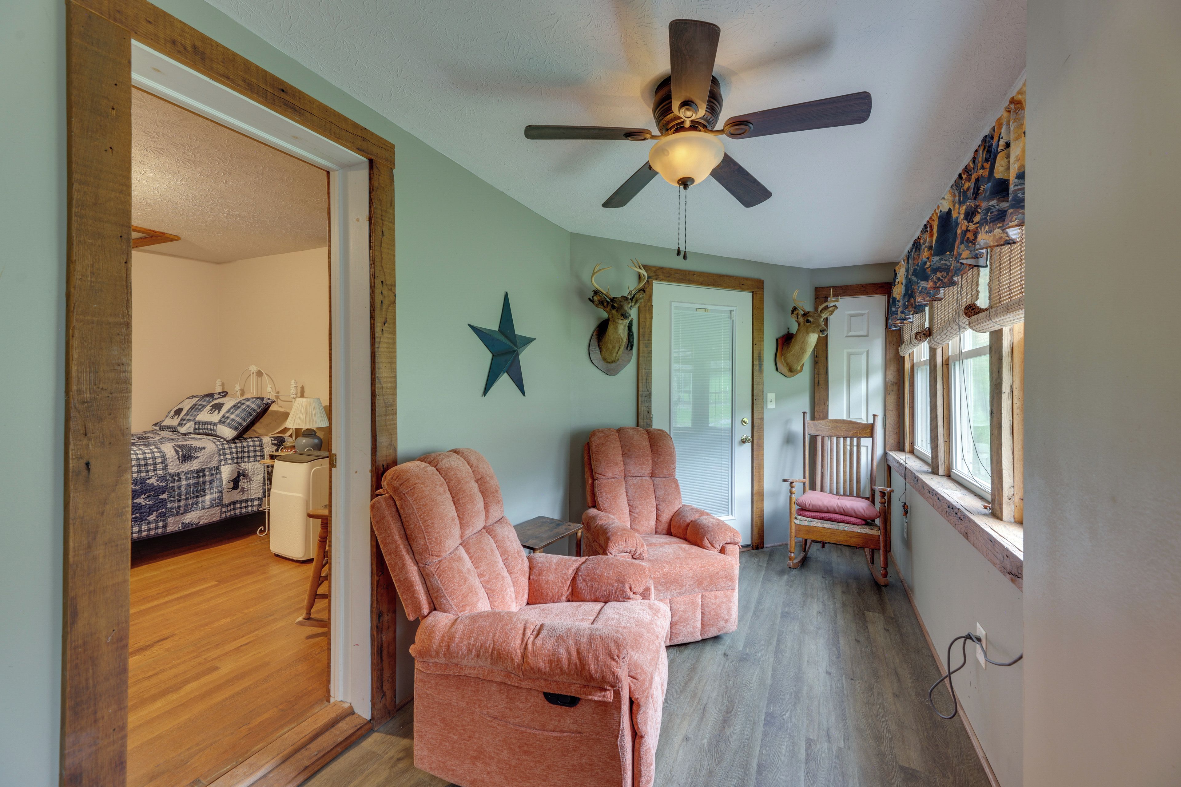 A room with a ceiling fan, a bed with a blue and white checkered bedspread, two pink armchairs, a rocking chair, and a star decoration on the wall.