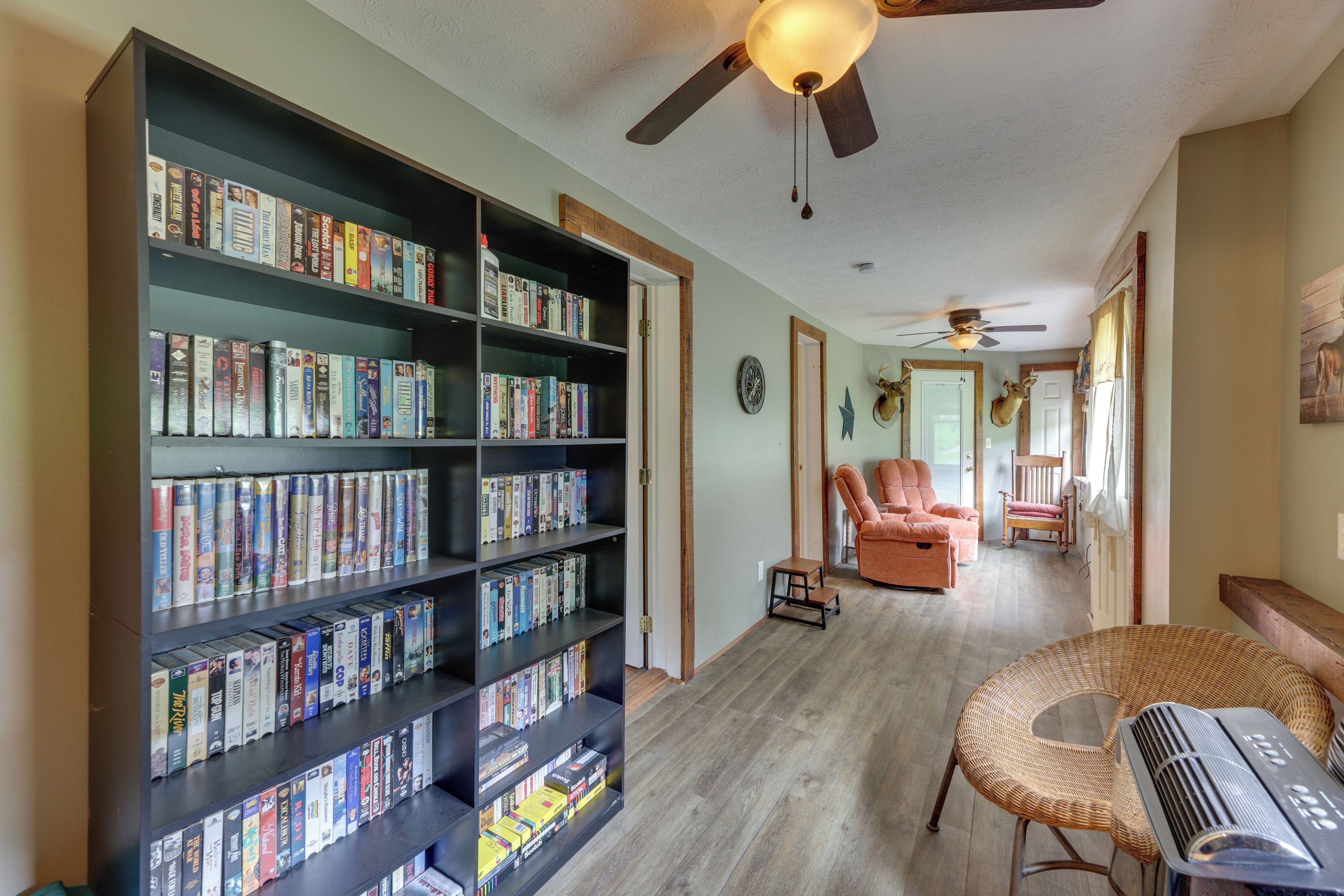 A room with a large bookshelf filled with books, a ceiling fan, a wicker chair, and a small table.