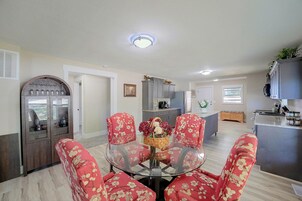 Uma área de refeições com mesa de vidro cercada por cadeiras de padrão floral, cozinha ao fundo e luminária no teto.