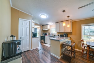 A kitchen with wooden flooring, white cabinets, a dining area with a table and chairs, and a view through the window.