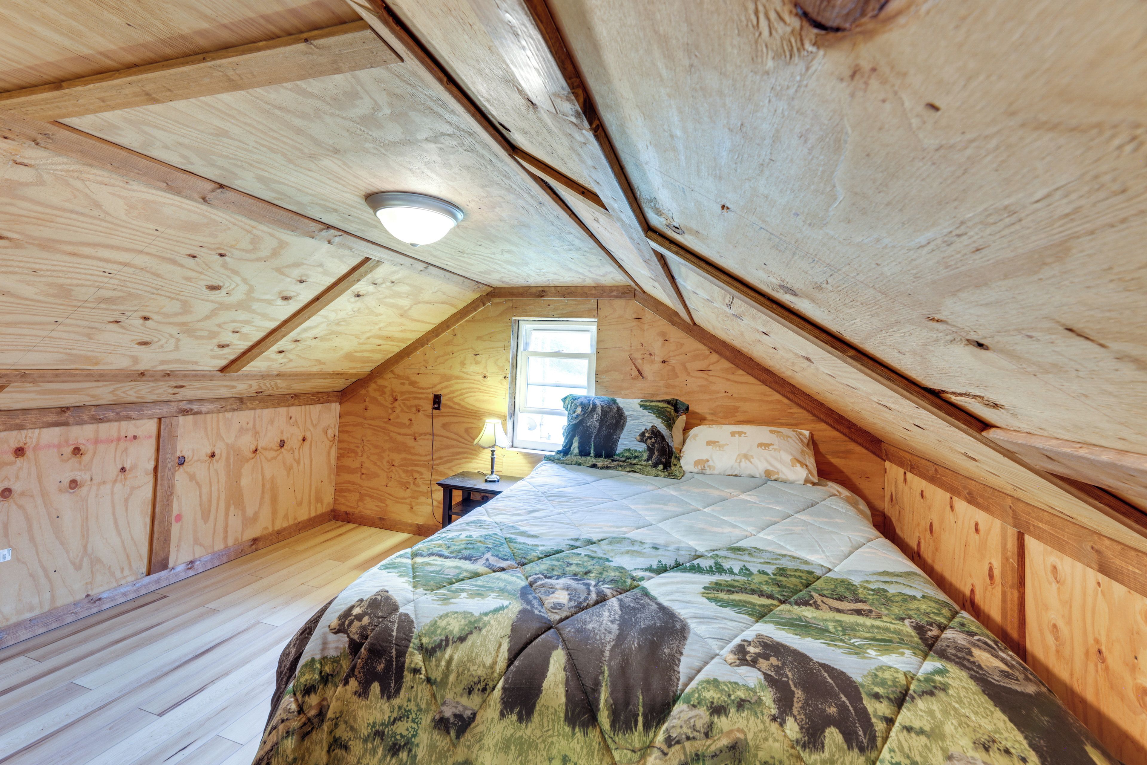 Un dormitorio con techo inclinado, paredes de madera, una cama con colcha de motivos naturales, una lámpara de noche y una ventana.
