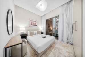 A modern hotel room with a large bed, bedside tables, a desk, and a view of the city through a sliding glass door. - Stayshort - Cosy 1 Bedroom in MBR City Sleeps 4 With Balcony (Dubai)