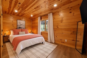 Ein Schlafzimmer mit Holzwänden, einem Bett, einem Fenster mit Vorhängen und einem an der Wand befestigten Fernseher.
