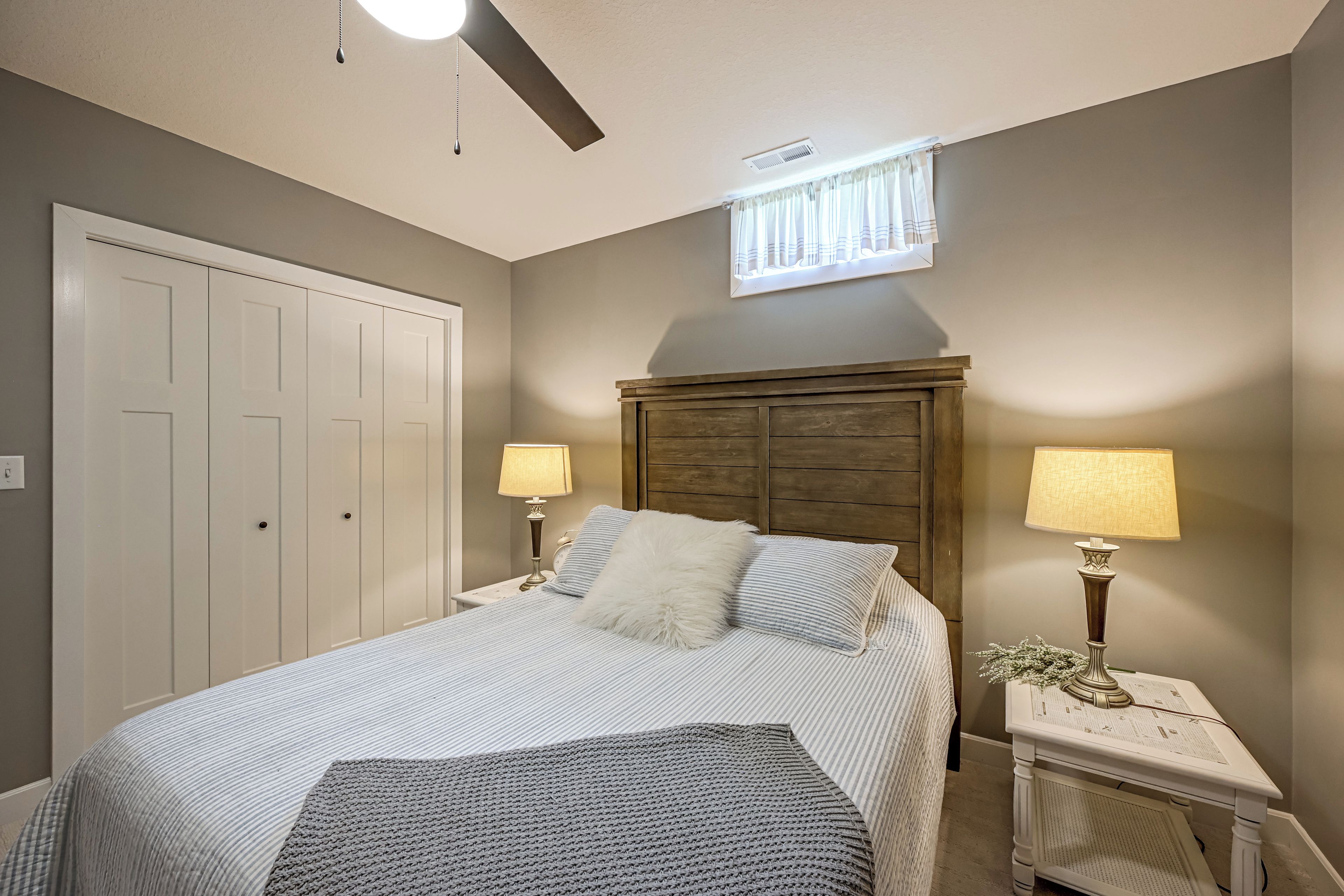 Un dormitorio con una cama, dos mesitas de noche con lámparas, un ventilador de techo y una ventana con cortinas.