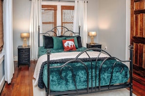 Un dormitorio con una cama de estructura metálica, dos mesitas de noche con lámparas, una ventana con cortinas y una puerta de madera.