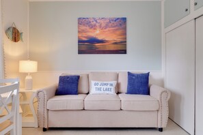 Een kamer met een beige bank, twee kussens, een wandlamp en een schilderij van een zonsondergang.