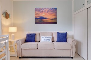 Una habitación con un sofá color beige, dos almohadas, una lámpara de pared y una pintura de atardecer.