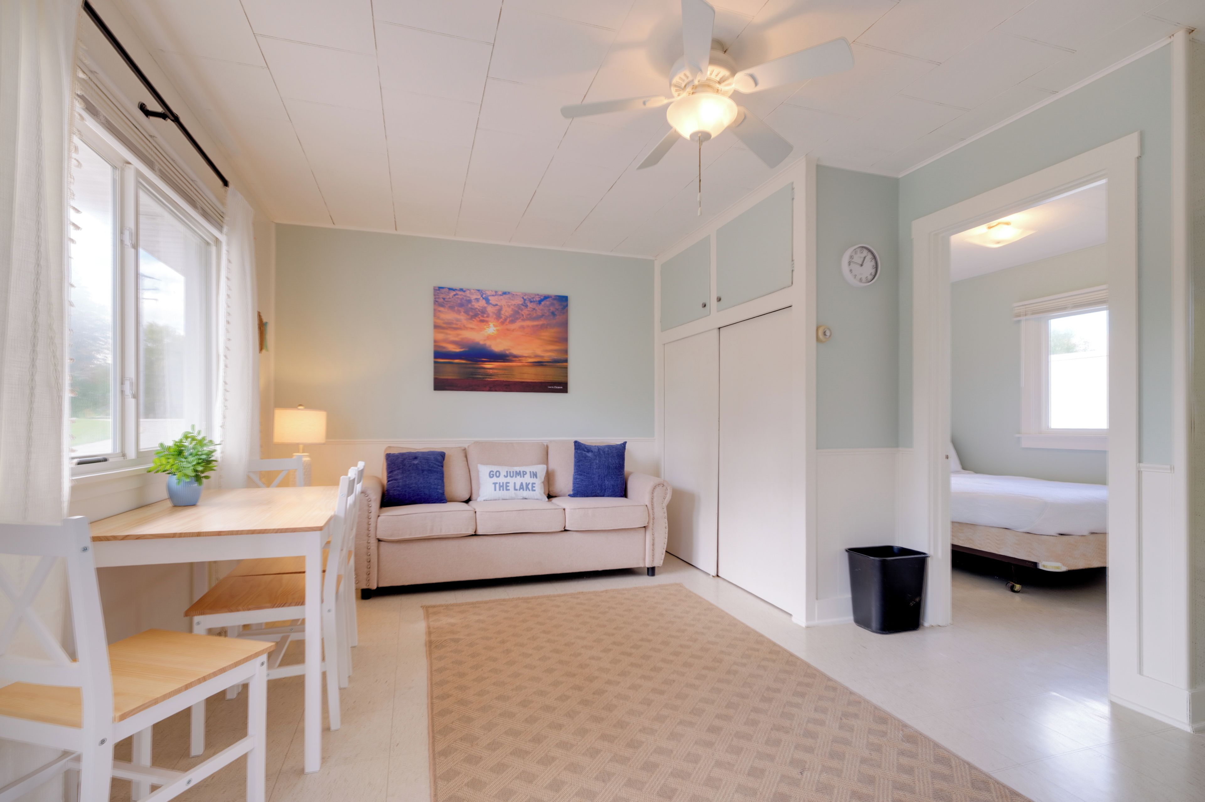 A room with a white ceiling fan, a beige sofa with blue cushions, a dining table with chairs, a small rug, and a wall clock.