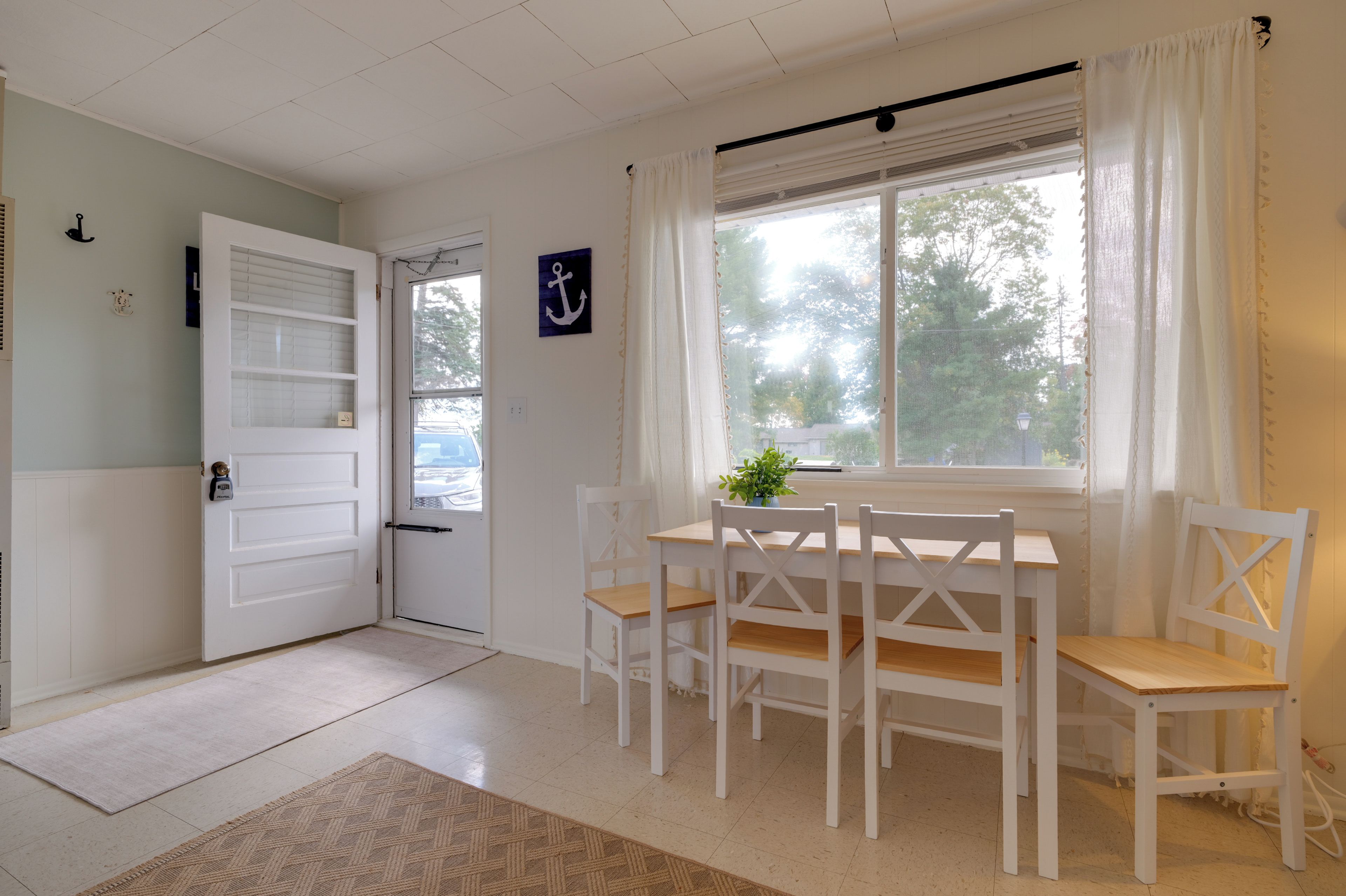 Una habitación con una puerta blanca, una ventana con cortinas, una mesa con sillas y una planta pequeña sobre la mesa.