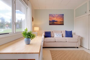 A room with a beige sofa, a blue cushion, a potted plant, a rug, and a sunset painting on the wall.