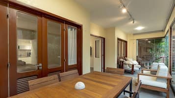 Un espace repas avec une table et des chaises en bois, une cuisine visible à travers une porte vitrée, et un coin salon avec un canapé et des fauteuils.