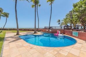Uma piscina de borda curva, rodeada por um deck ladrilhado, palmeiras e vista para o oceano.