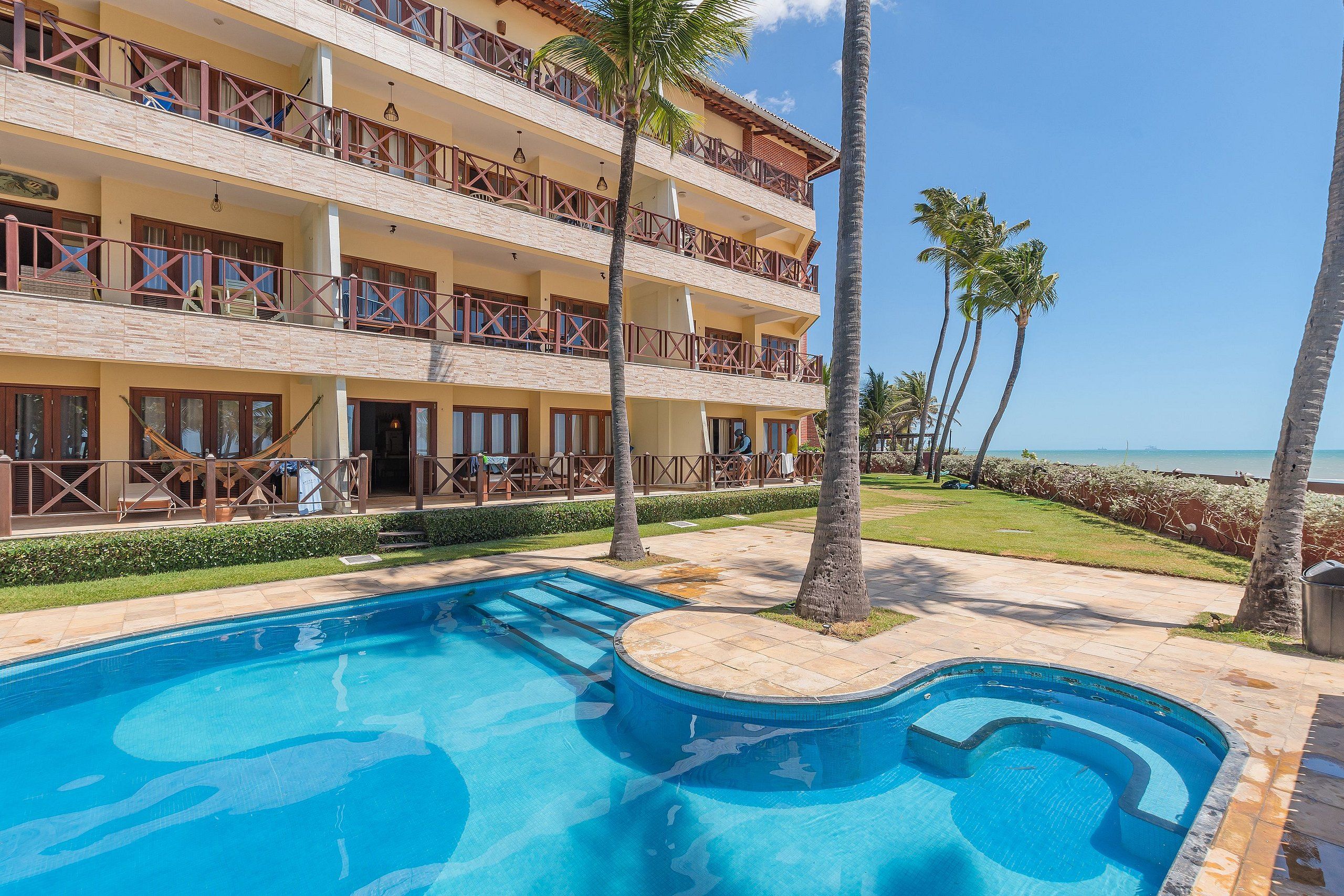 Hotel com vários andares, piscina, palmeiras e vista para o oceano.