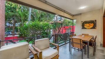 Un interior moderno con un balcón, una zona de comedor con mesa y sillas, y un ventanal que ofrece vistas a zonas verdes y un edificio rojo.