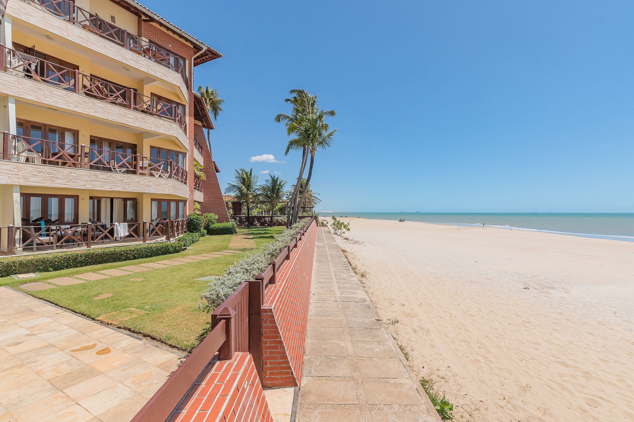 Un hotel frente a la playa con un largo paseo pavimentado que conduce a la arena, palmeras y una vista despejada del océano.