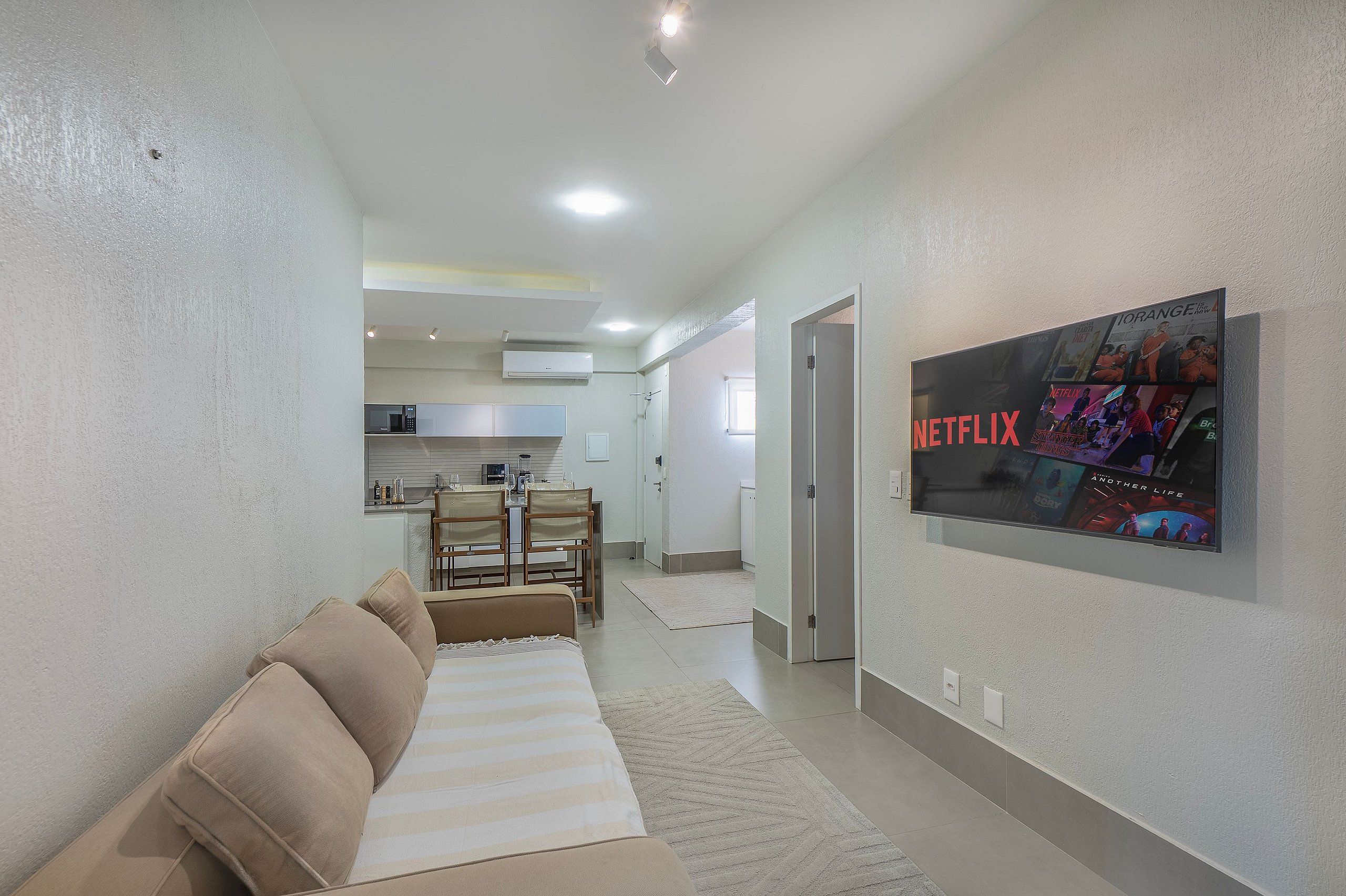 Una sala moderna con un sofá, un televisor de pantalla plana montado en la pared y una cocina al fondo.