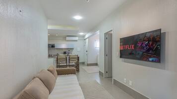 Una sala moderna con un sofá, un televisor de pantalla plana montado en la pared y una cocina al fondo.